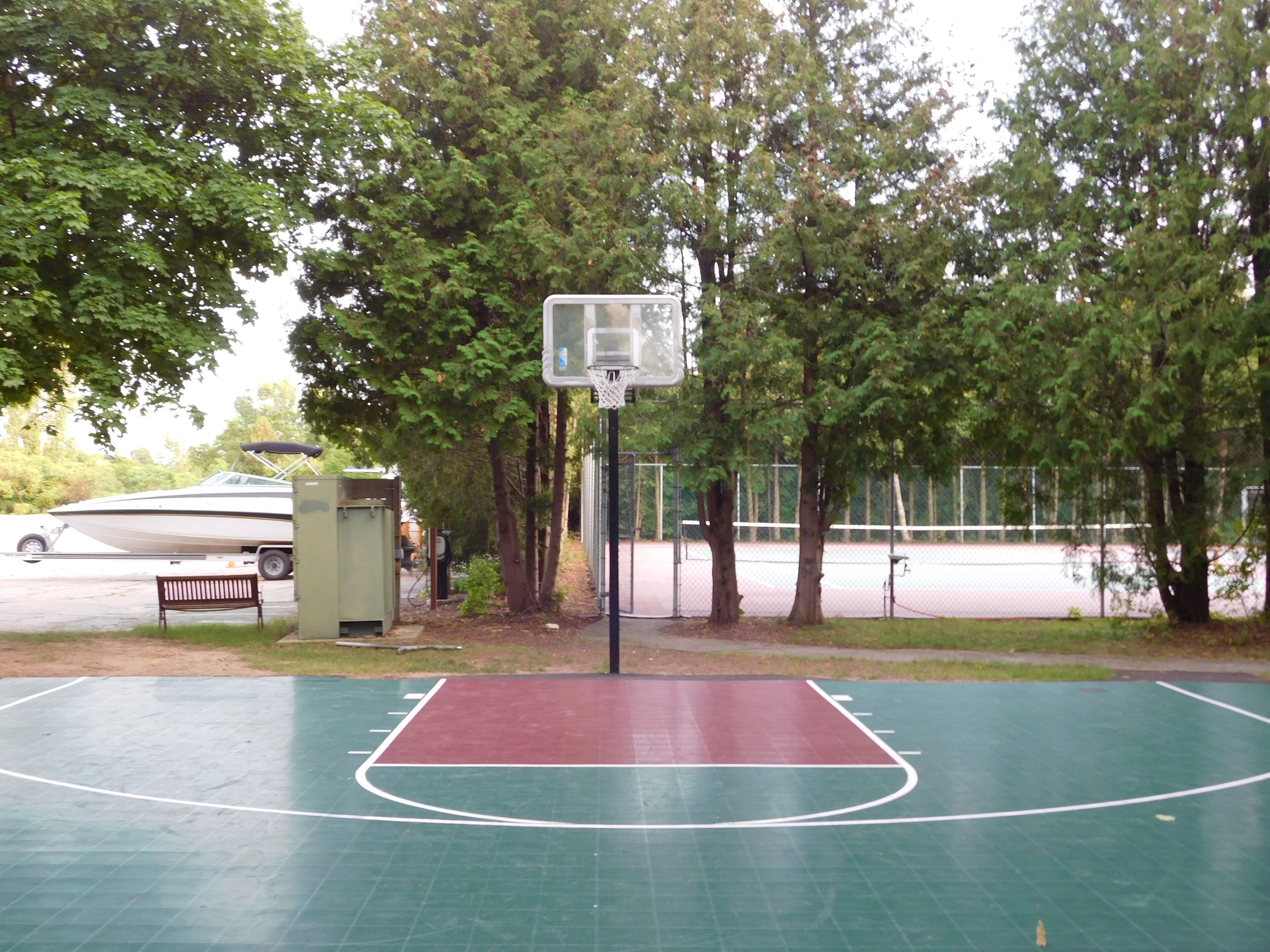Quadra de basquete ao ar livre com superfície verde e área de jogo vermelha, cercada por árvores e uma grade.