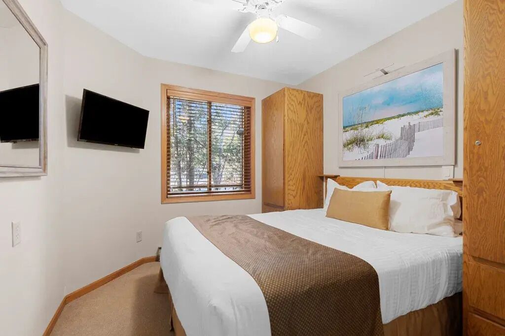A bedroom with a bed, a television, a window with blinds, a ceiling fan, and a painting on the wall.