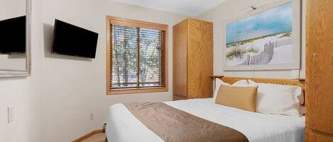 A bedroom with a bed, a television, a window with blinds, a ceiling fan, and a painting on the wall.