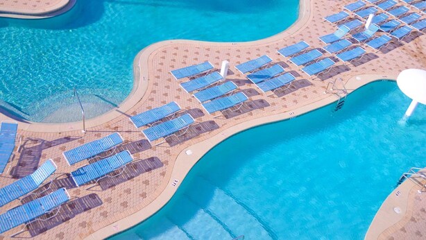 Vue aérienne d’un complexe hôtelier doté d’une grande piscine, de chaises longues et d’une plage de sable.