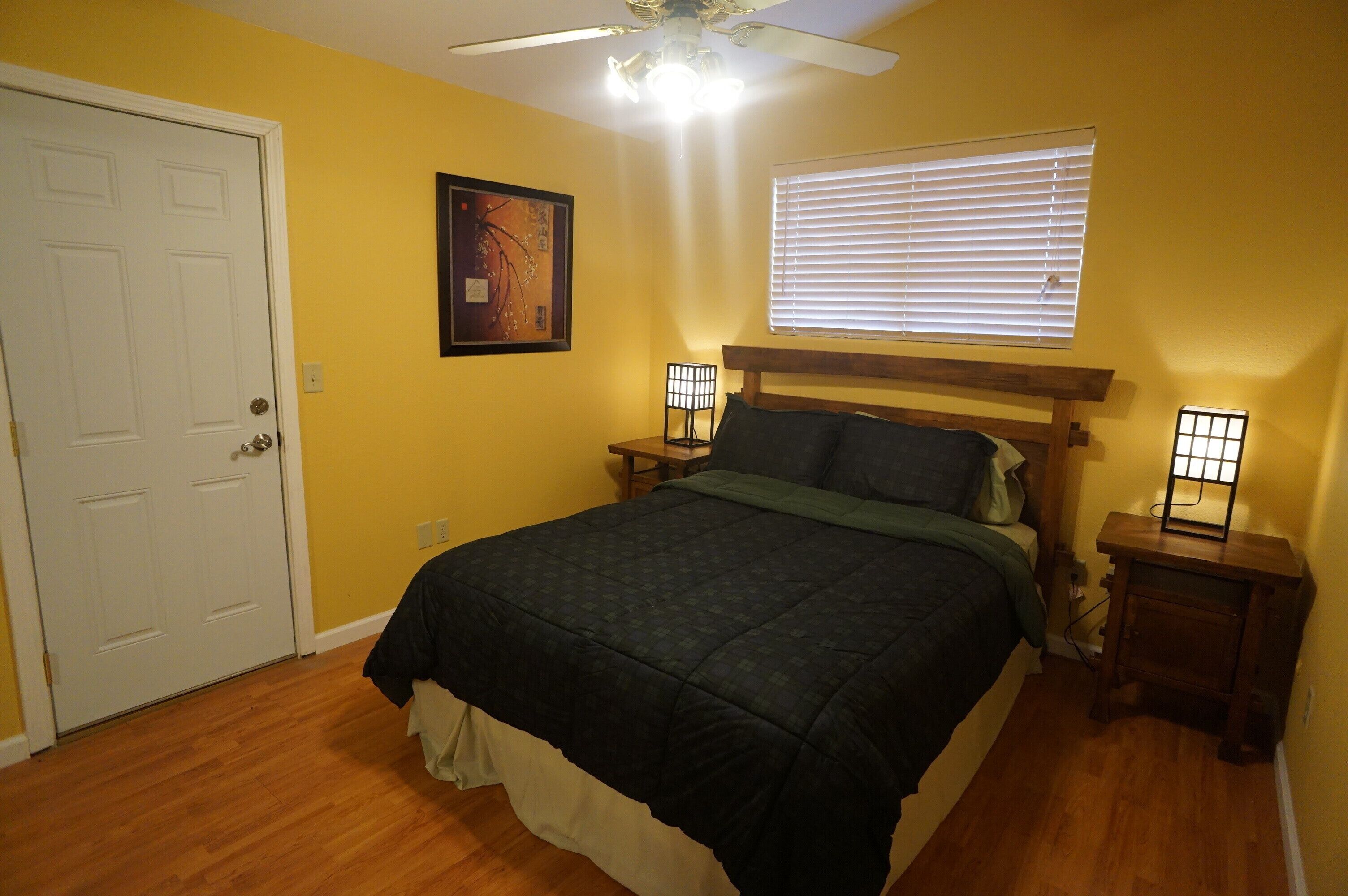 A bedroom with a bed, a ceiling fan, a window with blinds, and a door.