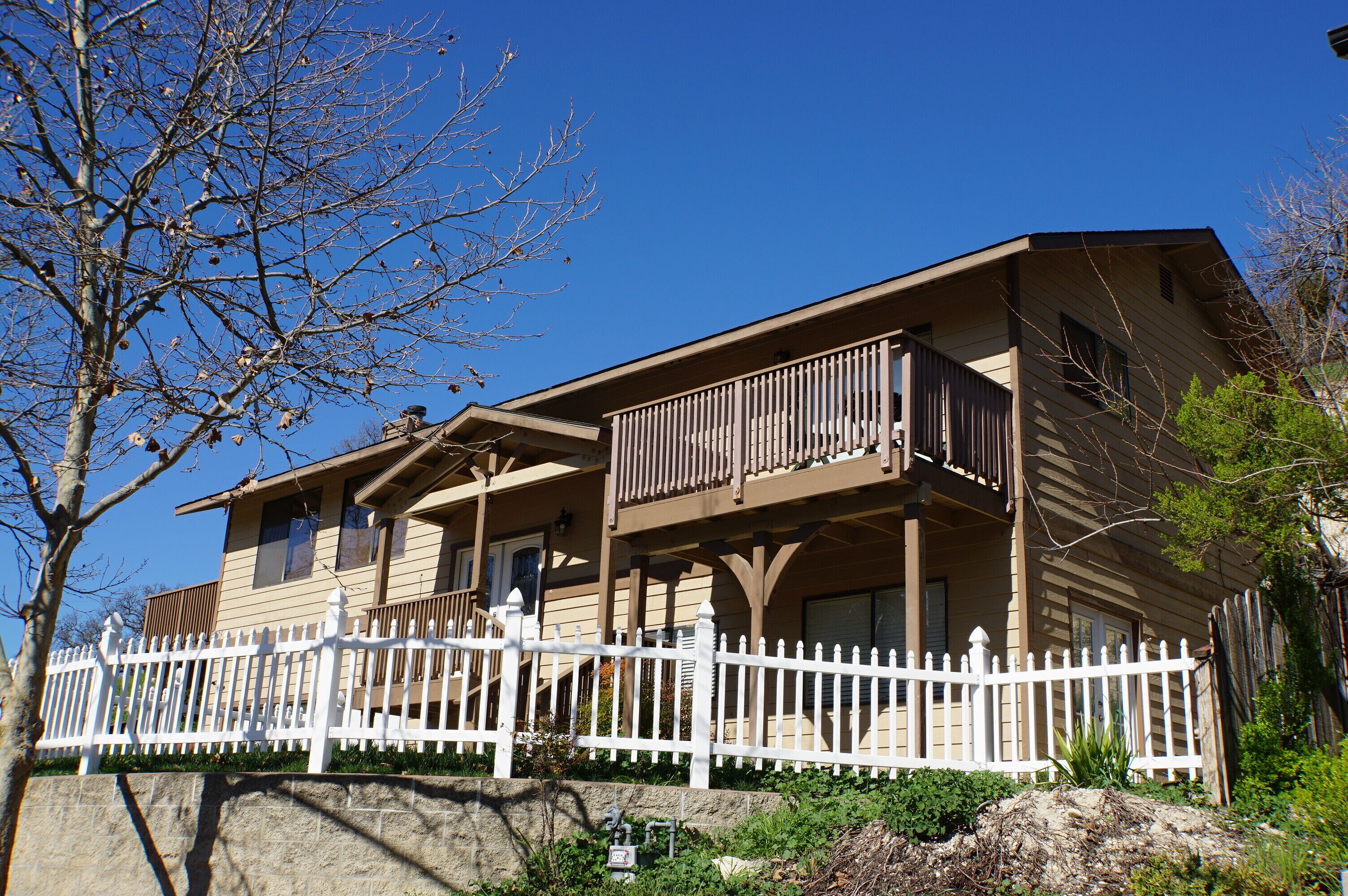 A two-story building with a white picket fence, a balcony on the second floor, and a stone wall in the foreground.