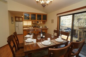 On aperçoit un coin repas avec une table en bois dressée pour quatre personnes, entourée de chaises en bois. Au fond, on distingue une cuisine avec des placards en bois et un réfrigérateur.