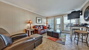 Una sala de estar con sofá, sillones, una mesa de centro y televisión. Hay una ventana con persianas, una chimenea y un comedor con mesa y sillas.
