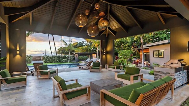 Un espace de détente extérieur avec des meubles en bois et des coussins verts, une piscine et une vue sur le jardin environnant.