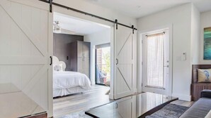 Un intérieur moderne avec une table basse en verre, un lit recouvert de linge de lit blanc et une porte coulissante de style rustique donnant sur une autre pièce.