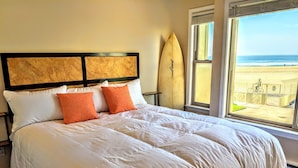 Ein Bett mit weißer Bettwäsche und orangefarbenen Kissen, ein Surfbrett, das an der Wand lehnt, und Blick auf den Strand durch ein Fenster.