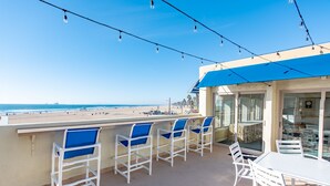 Ein Balkon mit weißen Stühlen und einem Tisch, der Blick auf einen Strand unter wolkenlosem Himmel bietet.