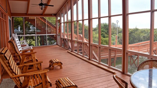 Een overdekt terras buiten met houten stoelen en tafels, grote ramen en uitzicht op een houten brug en groen.