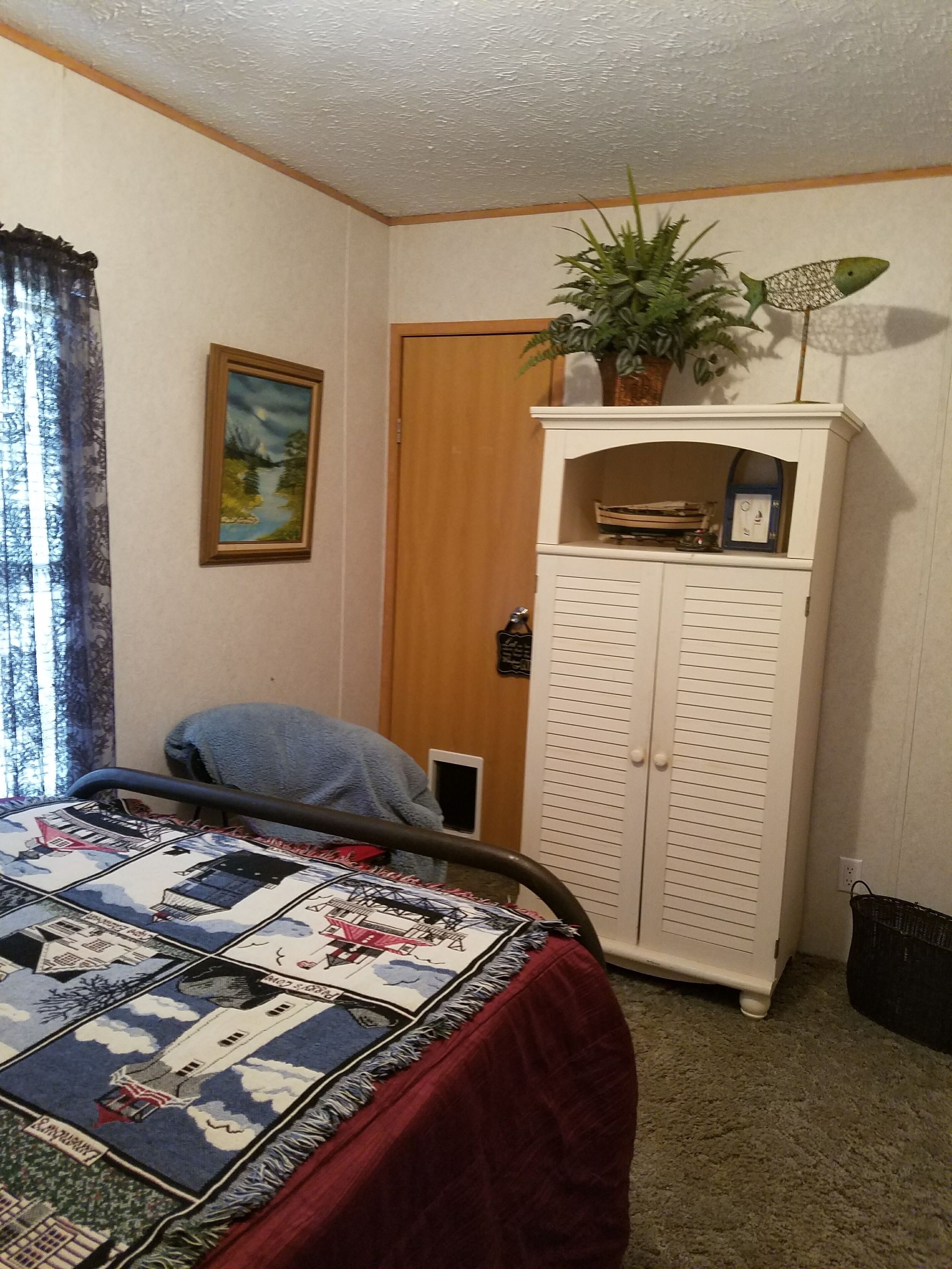 A bedroom with a bed, a painting, a plant, and a cabinet.