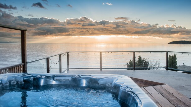 Un jacuzzi en una terraza de madera con vista a un cuerpo de agua y una puesta de sol al fondo.