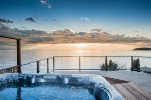 Een hottub op een houten terras met uitzicht op een wateroppervlak, met een zonsondergang op de achtergrond.