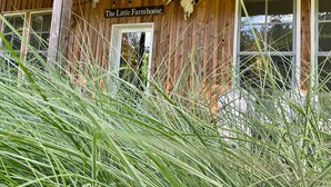 Una construcción de madera con un letrero que dice "The Little Farmhouse" y una puerta. Hay dos ventanas y una guirnalda de luces.