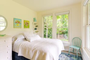 A bedroom with a bed, a dresser, a chair, and two windows offering a view of greenery.