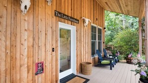 Fachada de madera de una propiedad llamada "The Little Farmhouse", con un porche cubierto, mobiliario exterior y un paisaje natural de fondo.