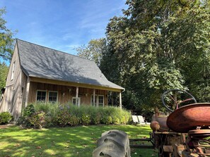 A rustic wooden house with a porch, surrounded by lush greenery and a large tree.