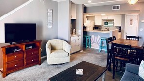 A living room with a TV, sofa, coffee table, and a chair. There's a staircase leading to a kitchen area with a bar and a dining table. - August Dates Still Available This Summer - Enjoy this Comfy 3 Bedroom Unit! (Orange Beach)