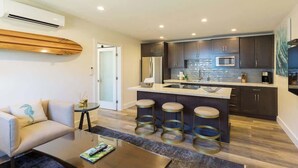 A modern kitchen with dark cabinets, a stainless steel refrigerator, and a microwave. There is a bar area with stools and a glass-top table. - Villa 4 (Kihei)
