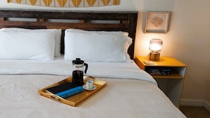 Una cama bien hecha con sábanas blancas, una bandeja con un libro, una cafetera y una taza sobre la cama.