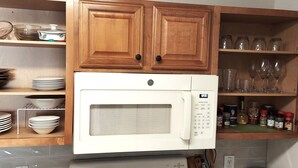 Una cocina con armarios de madera, un microondas blanco y estantes con vajilla, vasos y especias.