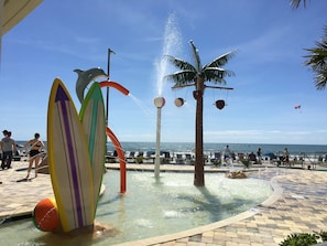 A water feature with a palm tree and surfboards, surrounded by a pool area with lounge chairs and a view of the ocean. - Spacious Ocean Front  Modern Decor.Spectacular View 2 Full Bath , 2nd floor (Myrtle Beach)