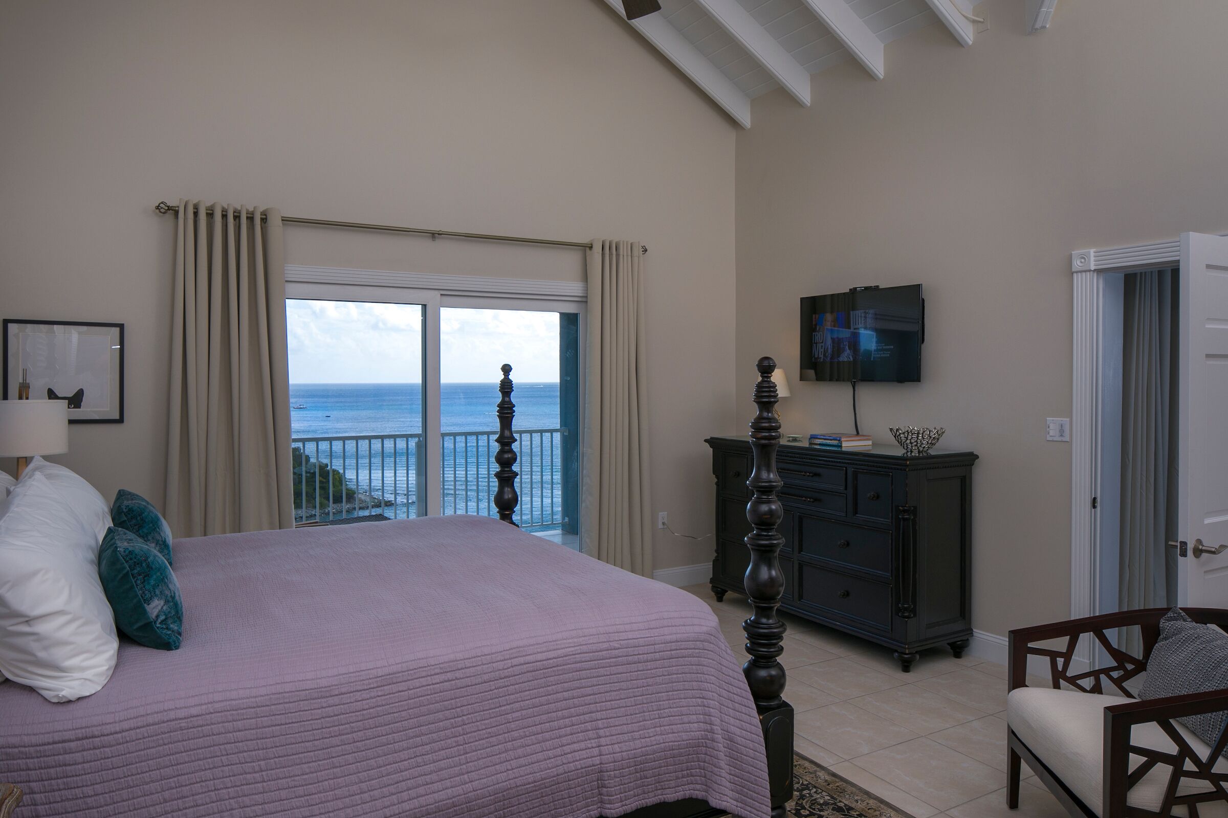 A bedroom with a large bed, a dresser, a television, and a view of the ocean.