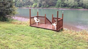 Une terrasse en bois avec deux chaises blanches donnant sur une rivière paisible, le tout entouré d’une forêt dense.