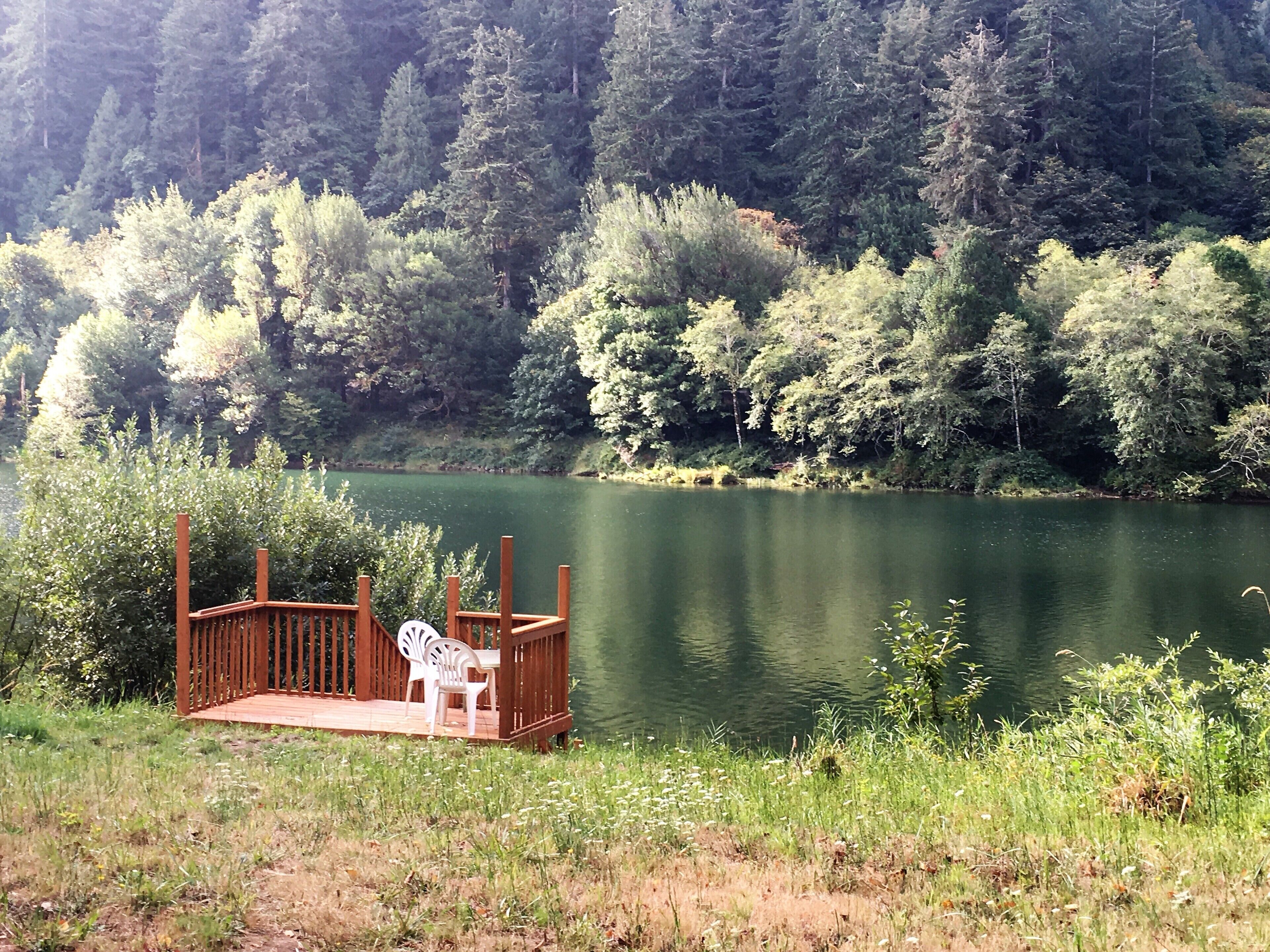 A wooden deck with two white plastic chairs and a table, overlooking a calm lake surrounded by dense forest.