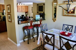 Un comedor con una mesa dispuesta para cuatro personas, rodeada de sillas con cojines rojos. En la mesa, una bandeja roja con un tarro y una vela. La habitación tiene armarios de madera y una cocina al fondo.
