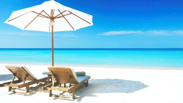 Una playa con dos sillas de madera y una sombrilla blanca.