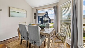 En matplats med ett träbord och stolar, utsikt över ett hus genom fönstret och en tavla på väggen.