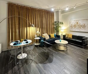 Een moderne woonkamer met een donker houten vloer, een ronde salontafel, een blauwe bank, een zwarte bank en een eethoek met een metalen stoel en een glazen tafel.