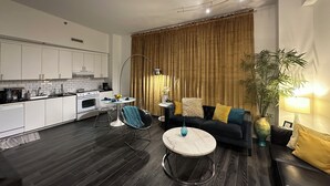 Un espace de vie compact comprenant un coin cuisine, un canapé, une table basse et une grande fenêtre avec des rideaux.
