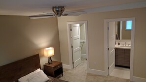 Un dormitorio con cama, mesita de noche, lámpara y ventilador de techo. Se ve un baño a través de una puerta abierta.