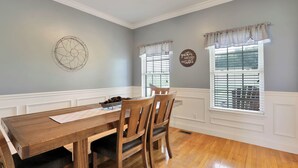 A dining room with a wooden table and chairs, a decorative wall piece, and two windows with valances. - Riverfront home on South Holston River Perfect for Fishing, Hiking, Relaxing (Bristol)