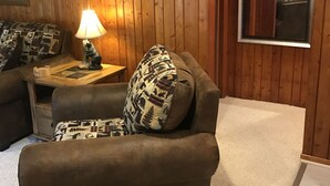 Un salon chaleureux aux murs lambrissés, avec un canapé marron agrémenté de coussins à motifs, une petite table sur laquelle est posée une lampe, et un grand miroir qui reflète la pièce.