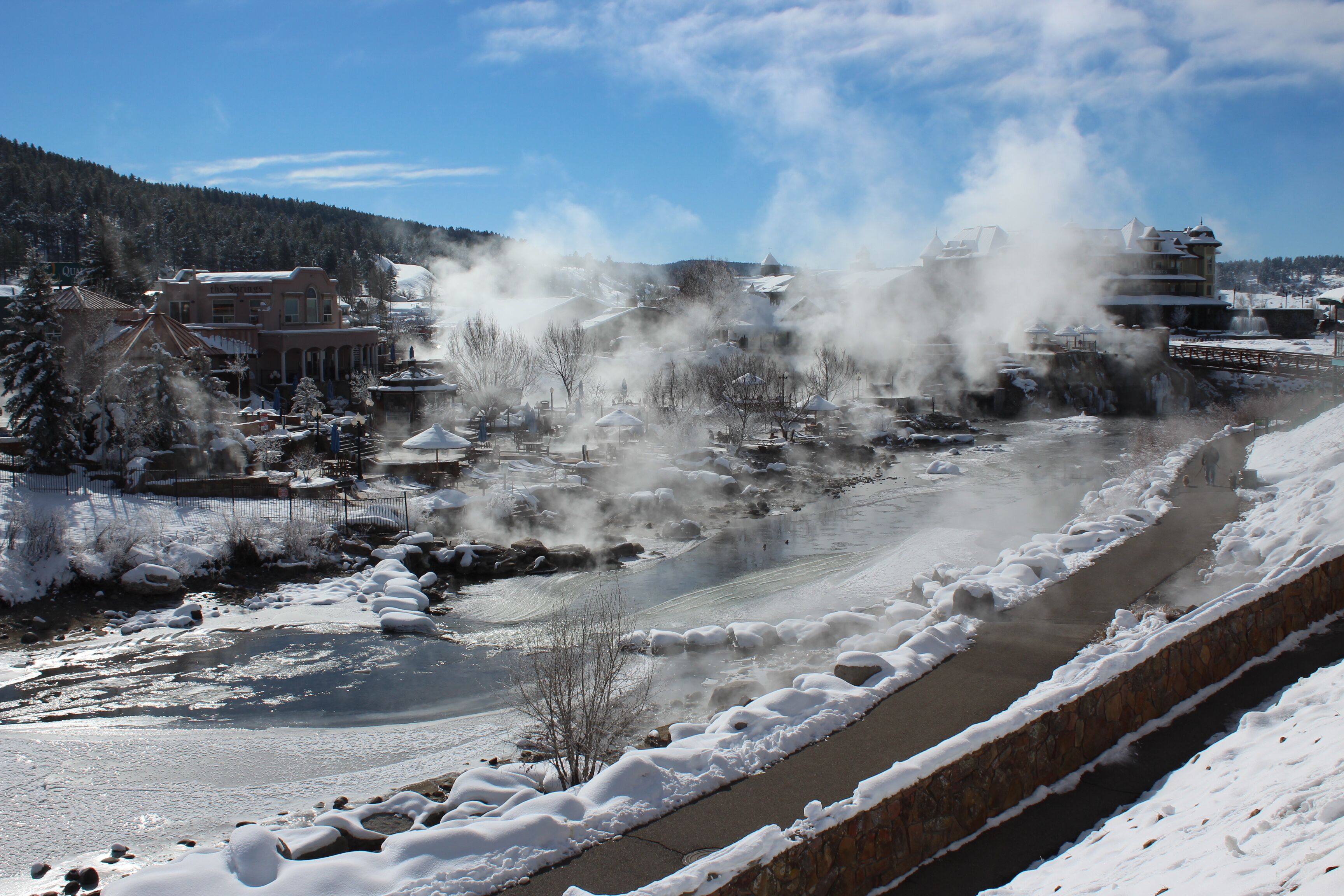 A snow-covered river with steam rising from the water, surrounded by buildings and trees.