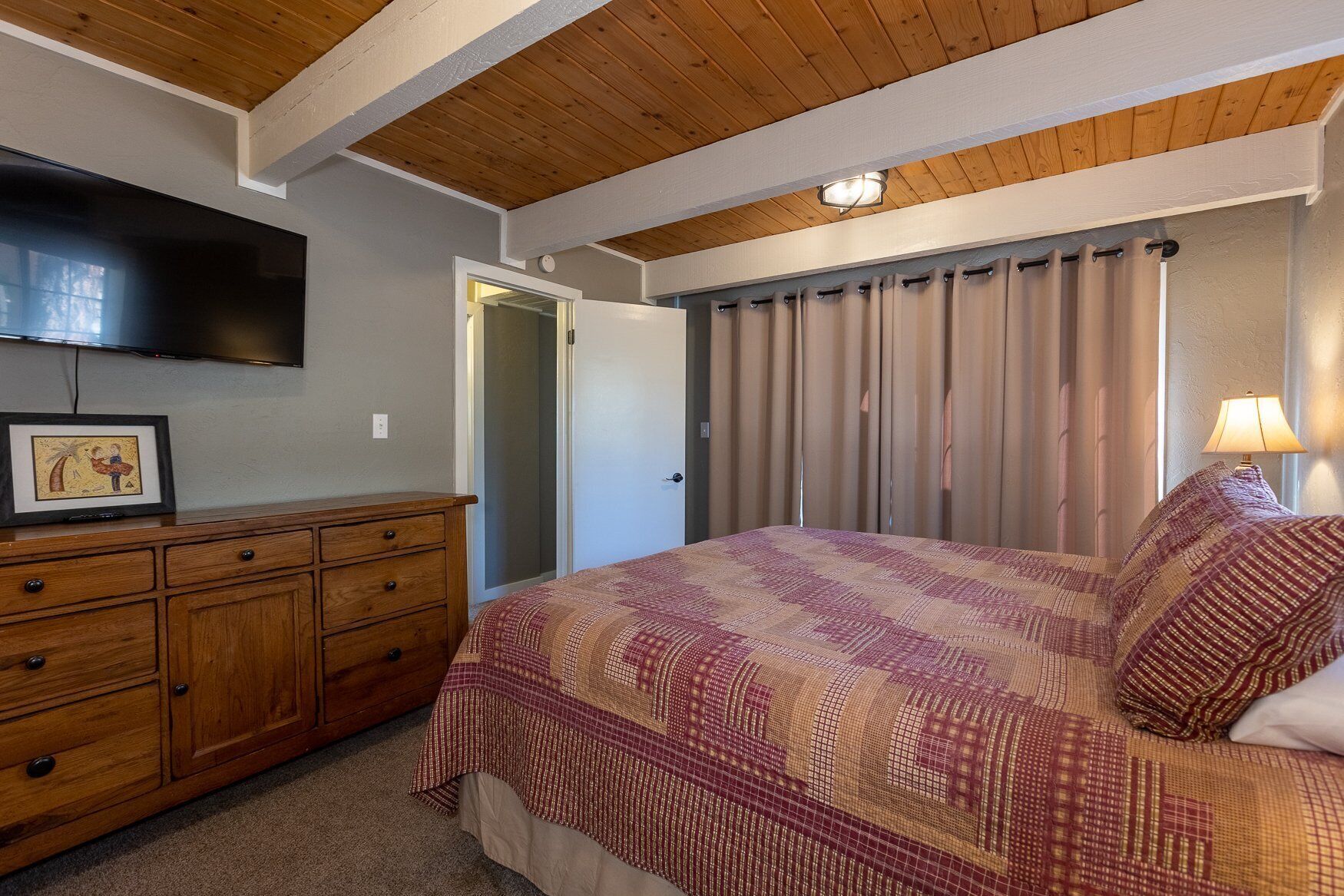 Un dormitorio con techo de madera, una cama con ropa de cama estampada, un aparador, un televisor y una puerta que da a otra habitación.