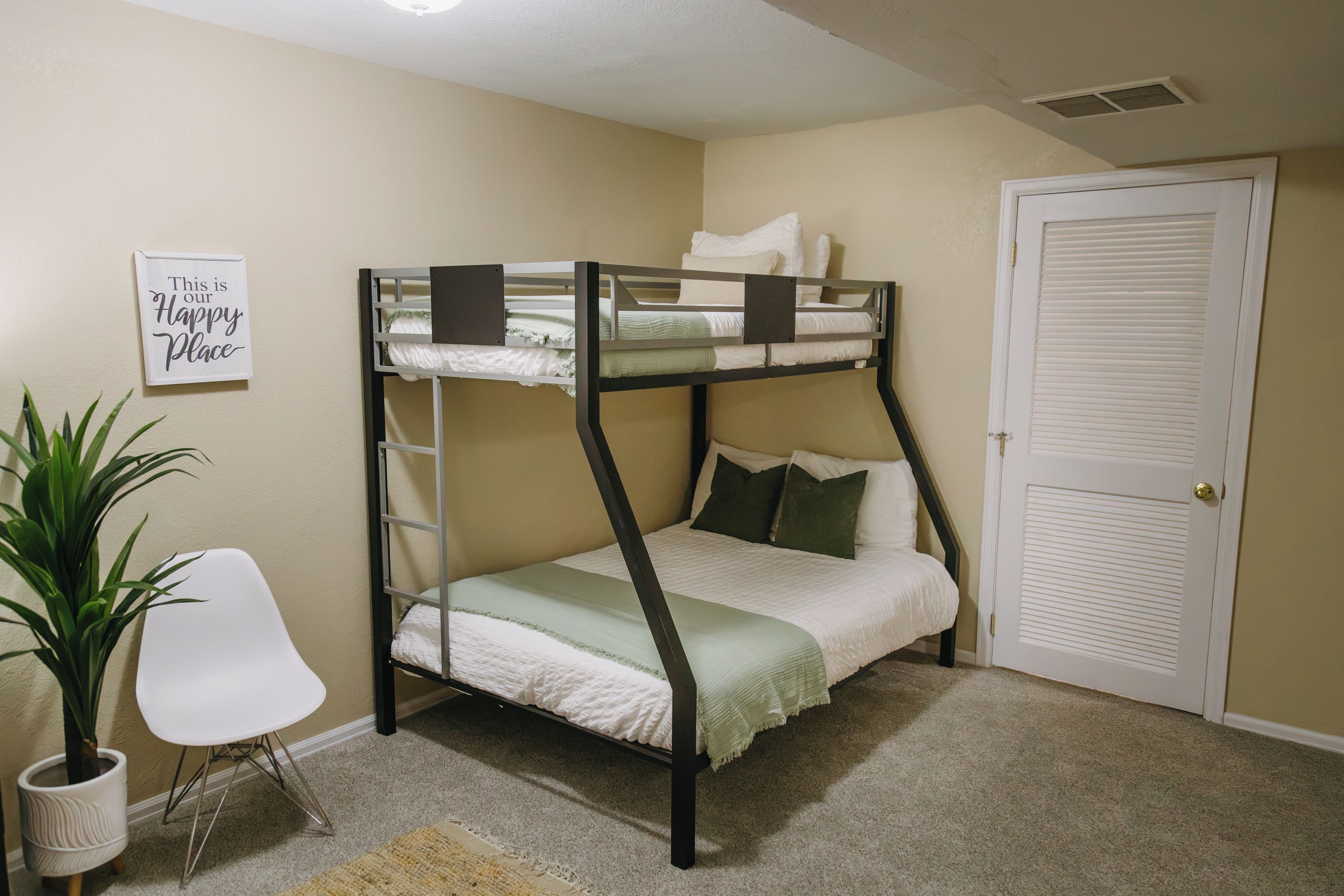 A bunk bed with a headboard, a white chair, a potted plant, and a door with a window.