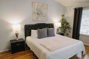 A bedroom with a large bed, two bedside tables with lamps, a wall-mounted artwork, a potted plant, and a window with curtains.