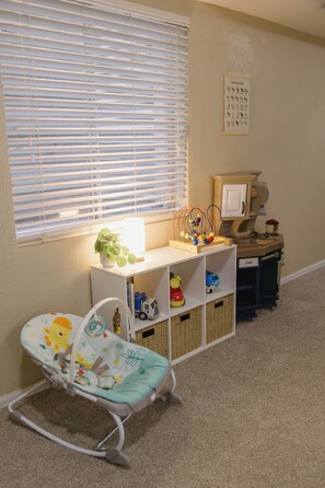 A room with a crib, a changing table, a shelf with toys, a window with blinds, and a wall calendar.