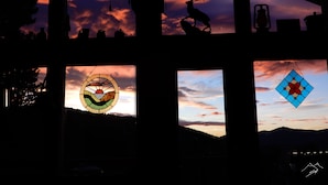 Ein Zimmer mit mehreren Fenstern, die verschiedene Ausblicke zeigen, darunter ein Sonnenuntergang, die Silhouette eines Rehs und ein Buntglasfenster.