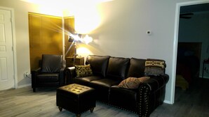 Una sala de estar con un sofá de cuero negro, un reposapiés a juego y una lámpara. Se observa una silla y un termostato instalado en la pared.