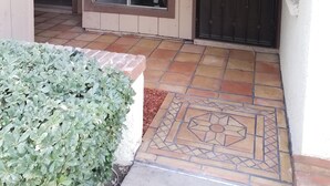 Un porche cubierto con piso de baldosas, un banco de madera, una ventana con persianas y una puerta metálica oscura.