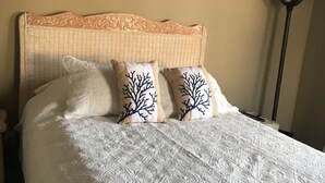 Cama com cabeceira trançada, roupa de cama branca e almofadas decorativas com estampas de árvores.