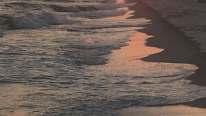 Praia ao pôr do sol, com ondas e algumas pessoas ao longe.
