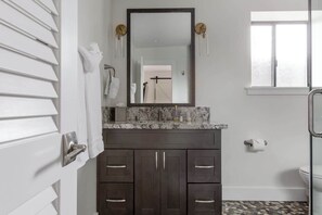 A bathroom with a dark wood vanity, a marble countertop, a large mirror, and a toilet paper holder. - Villa 8 (Kihei)