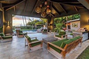 An outdoor lounge area with wooden furniture and green cushions, a pool, and a view of the surrounding greenery. - Villa 8 (Kihei)