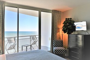 A hotel room with a balcony, a bed, a desk, a chair, a dresser, and a plant.