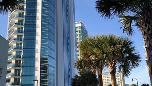 A tall, modern hotel with a glass facade and multiple balconies, surrounded by palm trees and a clear blue sky.